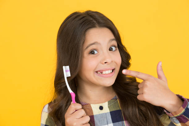 Young girl smiling, pointing at her teeth and holding a toothbrush in front of a yellow background