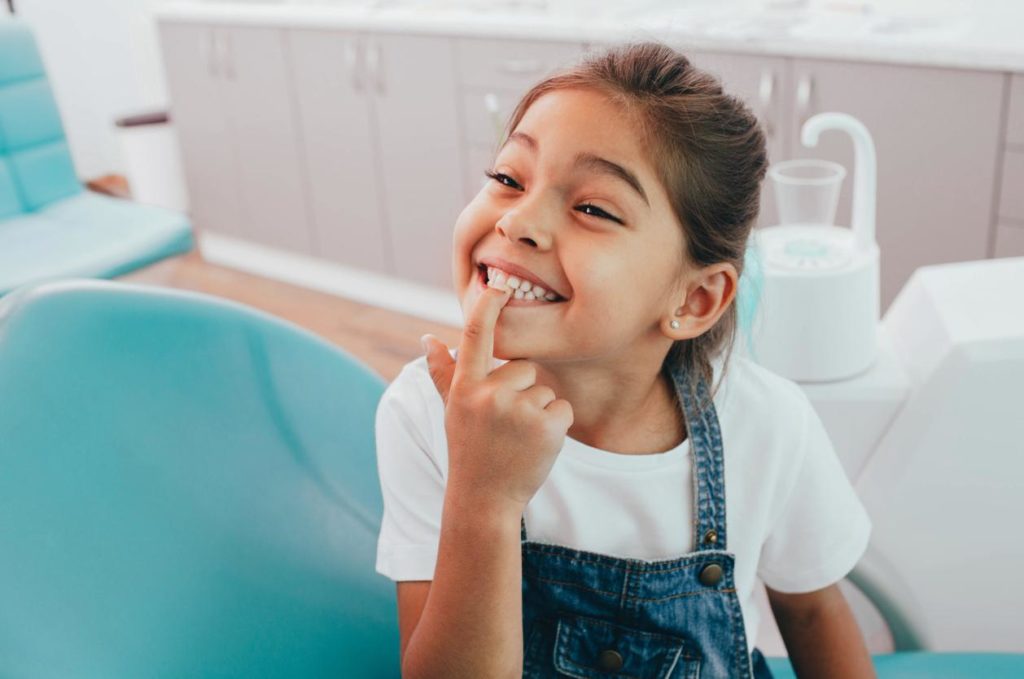 A child smiling in a blue dentist chair and pointing to her teeth.