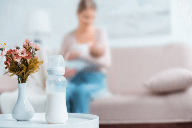 A baby bottle filled with milk sits on a white table beside a vase of flowers while a mother and baby sit on a couch in the background.