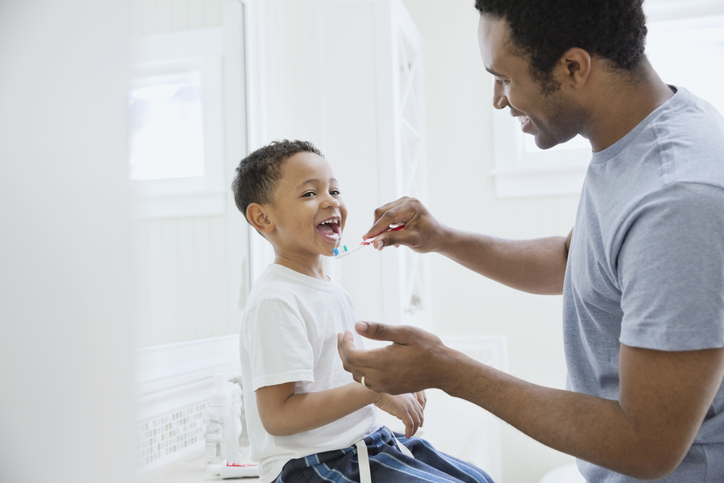 A father happily brushes his son's teeth in order to prevent cavities.