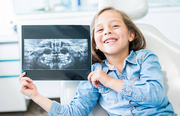 A young girl holding up a dental X-ray image while sitting in a dental chair.