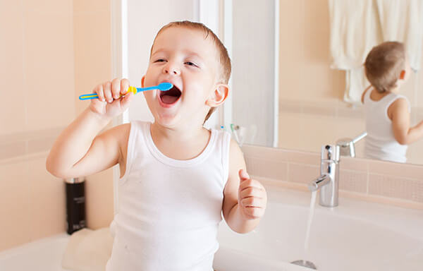 A young boy sitting at a bathroom sink and holding a toothbrush up to his mouth.