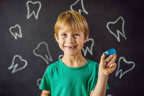 A young boy smiling and holding up a blue mouthguard as he stands in front of a blackboard with drawings of teeth on it.