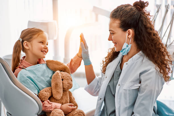A young girl sitting in a dental chair holding a stuffed animal gives a dentist a high-five.