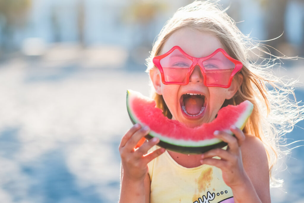 Young girl smiling while holding a slice of watermelon and wearing star-shaped pink sunglasses.